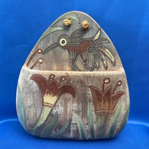 May include: A wooden, hand-painted decorative box with a hummingbird and floral design. The box features a brown hummingbird with yellow and brown accents, and two flowers with green leaves. The background is a weathered gray.