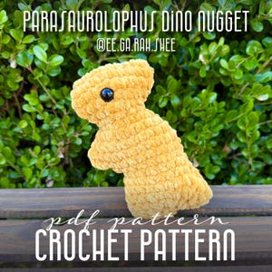 May include: A plush, yellow Parasaurolophus Dino Nugget, with a black eye, is displayed against a backdrop of green foliage and a wooden surface. The text "PARASAUROLOPHUS DINO NUGGET" and "pdf pattern CROCHET PATTERN" are visible.