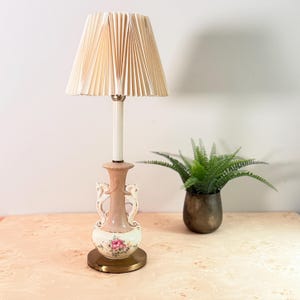 May include: A table lamp featuring a cream-colored pleated lampshade and a ceramic base. The base is shaped like an urn with two horse-head handles, finished in a soft peach and cream glaze, and decorated with a floral pattern of pink roses.