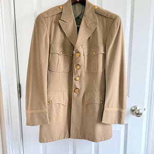 May include: Tan military-style jacket with gold buttons and four front pockets. The jacket has a notched collar and is hanging on a wooden hanger. The sleeves have a tan stripe detail near the cuffs. The jacket is a neutral color.