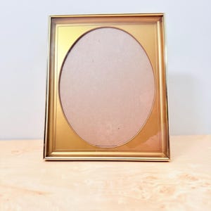 May include: A rectangular gold-toned picture frame featuring a central oval-shaped opening. The frame has a smooth, metallic finish with a thin border surrounding the oval cutout, designed to hold a photograph or artwork within its structure.