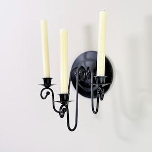 May include: Black metal wall sconce with three candle holders, each holding a cream-colored candle. The sconce has a circular backplate and decorative, curved arms. The candles are unlit.