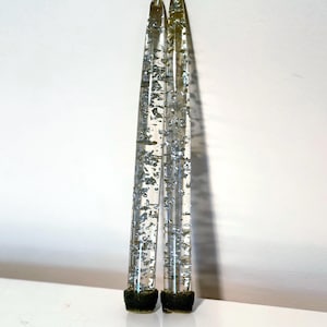 May include: Two clear taper candles with silver glitter suspended inside. The candles have a dark green base and silver wicks. The candles are standing upright against a white background.