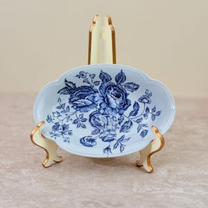 May include: An oval ceramic dish featuring a blue floral pattern on a light blue background. The dish is displayed on a cream-colored stand with gold-toned edges, showcasing detailed illustrations of roses and leaves across the surface.