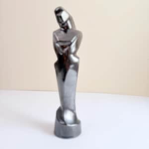May include: A tall, slender, abstract sculpture with a smooth, reflective dark metallic finish. The figure features a rounded base, a narrow waist, and a stylized, elongated upper section that curves slightly forward, creating a continuous form.