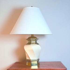 May include: A cream-colored table lamp with a white lampshade. The lamp base has a gold-colored metal trim and a twisted design. The lampshade is a classic cone shape. The lamp is sitting on a wooden table.
