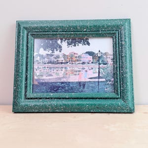 May include: A rectangular picture frame with a textured, speckled dark green finish. The frame contains a print depicting a waterfront scene with houses, palm trees, a street lamp, and two figures standing near the water's edge under a tree.