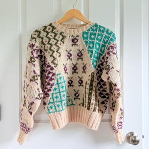 May include: A long-sleeve knit sweater featuring a cream base color with a multi-colored geometric pattern in shades of teal, dark green, and purple. The garment has a ribbed crew neckline, ribbed cuffs, and a ribbed hem, all in a solid cream color.