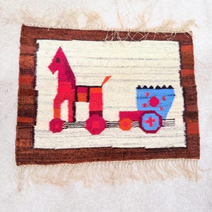 May include: A rectangular woven wall hanging featuring a central graphic of a red horse pulling a blue cart on wheels. The design is set against an off-white background with a dark brown border and light beige fringe along the top and bottom edges.