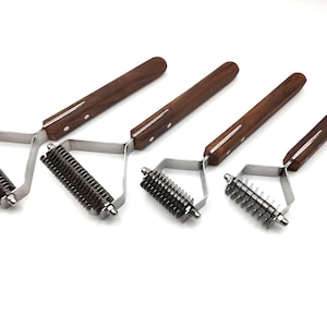 May include: Four pet grooming tools with wooden handles and metal blades are arranged on a white surface. The tools vary in size, with the largest on the left and the smallest on the right. The handles are a dark brown color.