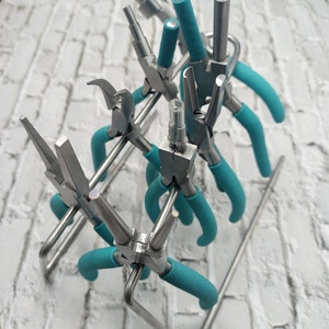 May include: A collection of jewelry pliers with teal handles, displayed in a silver metal stand. The pliers have various head shapes for different tasks. The background is a white brick wall.