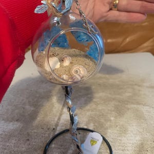 May include: A clear glass terrarium with sand, seashells, and blue accents. The terrarium is suspended from a decorative metal stand with a circular base. A starfish charm hangs from the top. The overall theme is coastal decor.