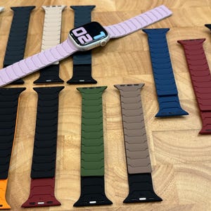 May include: A collection of silicone watch bands in various colors including white, black, beige, navy blue, olive green, tan, red, and orange, with one light purple band attached to a digital smartwatch displaying the time 20:02 on its screen.