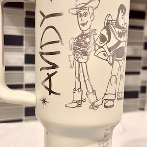 May include: Cream-coloured travel mug with illustrations of Woody and Buzz Lightyear from Toy Story, and other characters. The word "ANDY" is printed on the side. The mug has a white handle and lid.