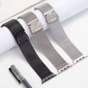 Könnte beinhalten: Drei Metall-Mesh-Uhrenarmbänder in Schwarz, Silber und Grau. Jedes Armband verfügt über einen einzigartigen Verschluss und Anschluss für eine Smartwatch. Ein Stift befindet sich im Vordergrund. Die Armbänder sind für einen modernen, eleganten Look konzipiert.