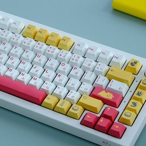 May include: A white keyboard with yellow and pink keycaps featuring Japanese characters and symbols. The keyboard has a white and pink wrist rest and a round knob on the right side.