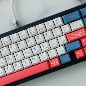 May include: A white and blue mechanical keyboard with Japanese characters on the keys. The keyboard has a red and white space bar and a black frame. The keyboard is connected to a white cable.