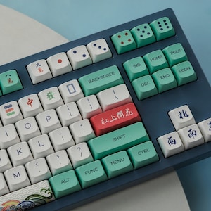Op de afbeelding: Een blauw en groen mechanisch toetsenbord met witte toetsen en Chinese karakters. Het toetsenbord heeft een verscheidenheid aan toetsen, waaronder functietoetsen, pijltoetsen en cijfertoetsen. Het toetsenbord heeft ook een rode toets met de Chinese karakters voor "杠上開花" (gang shang kai hua), wat "bloeien op de bar" betekent.