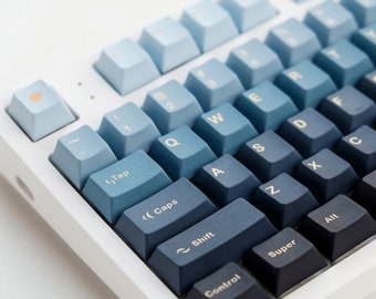 May include: A white mechanical keyboard with blue and gray gradient keycaps. The keys are labeled with letters, numbers, and symbols. The keyboard has a white frame and a white base.