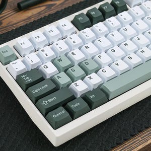 May include: A white and green mechanical keyboard with white keycaps and green accent keys. The keyboard has a variety of function keys and letter keys. The keyboard is on a black surface.