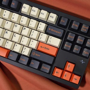 May include: A black mechanical keyboard with off-white and orange keycaps. The keyboard has a variety of keys, including function keys, number keys, and arrow keys. The keyboard is on a reddish-orange surface.
