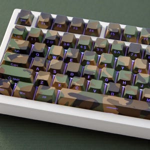 May include: A white mechanical keyboard with keycaps in a camouflage pattern of green, brown, and tan. The keyboard has a standard QWERTY layout with various function keys and symbols. The keys are illuminated with a purple backlight.