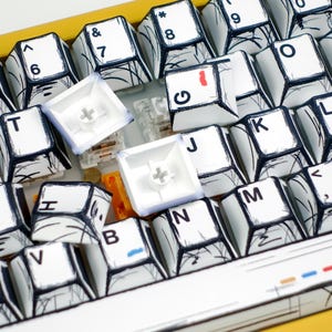 May include: Close-up of a mechanical keyboard with white keycaps featuring black line art and various letters and symbols. Several keycaps are removed, revealing the internal components. The keyboard has a yellow frame.