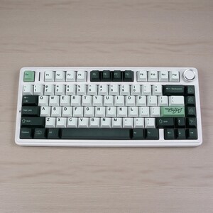 May include: A white mechanical keyboard with black and green accents. The keyboard features white keycaps with black lettering and green function keys. A green leaf design is on the right side. The keyboard sits on a light wood surface.
