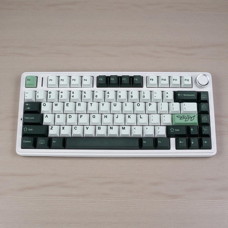 May include: A white mechanical keyboard with black and green accents. The keyboard features white keycaps with black lettering and green function keys. A green leaf design is on the right side. The keyboard sits on a light wood surface.