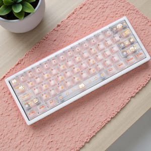 Könnte beinhalten: Eine weiße und rosa mechanische Tastatur mit transparenten Tastenkappen mit orangefarbenen Pfotenabdrücken und Katzenillustrationen. Die Tastatur liegt auf einer rosa strukturierten Oberfläche, mit einer kleinen Topfpflanze im Hintergrund.