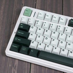 May include: A white and dark green mechanical keyboard with white keycaps and dark green accent keys. The keyboard has a minimalist design with a green Esc key and a lotus flower symbol on the space bar.