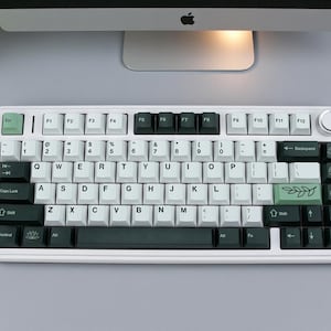 May include: A white mechanical keyboard with black and green keycaps. The keyboard has a standard QWERTY layout with a few custom keys, including a green Escape key and a green key with a leaf design. The keyboard is on a gray surface.