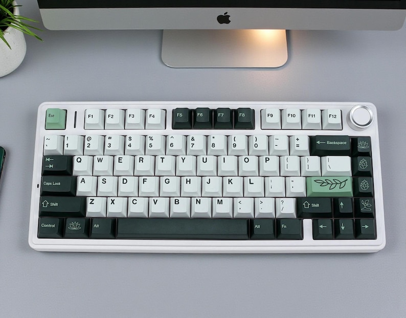May include: A white mechanical keyboard with black and green keycaps. The keyboard has a standard QWERTY layout with a few custom keys, including a green Escape key and a green key with a leaf design. The keyboard is on a gray surface.