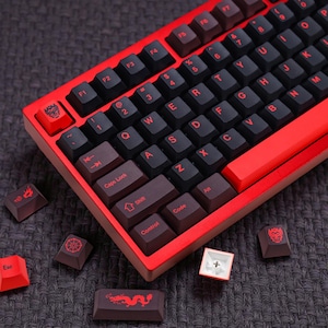 May include: A red and black mechanical keyboard with custom keycaps. The keyboard has a red frame and black keys with red lettering. Several custom keycaps with unique designs are scattered around the keyboard.