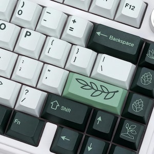 May include: Close-up of a mechanical keyboard with white and dark green keycaps. The keyboard features a variety of symbols and letters, including the word "Backspace" and the word "Shift". A light green keycap has a leaf design.