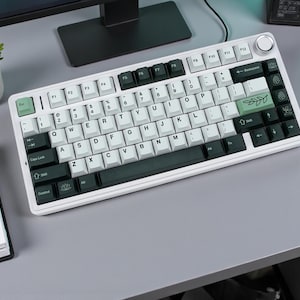 May include: A white mechanical keyboard with black and white keycaps. The keyboard has a silver frame and a volume control knob. Some keys are green with leaf designs. The keyboard is on a gray surface.