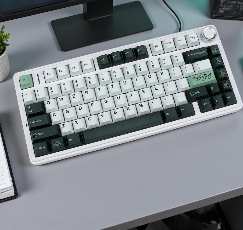 May include: A white mechanical keyboard with black and white keycaps. The keyboard has a silver frame and a volume control knob. Some keys are green with leaf designs. The keyboard is on a gray surface.