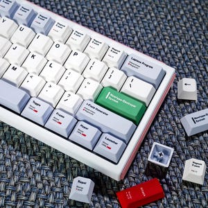 May include: A white and pink mechanical keyboard with various keycaps. The keyboard has white, gray, and green keys, with text such as "Newtype-Destroyer System" and "Attack". Several loose keycaps are scattered around the keyboard.