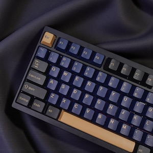 Puede incluir: Un teclado mecánico con marco azul oscuro y teclas azul claro. El teclado tiene teclas de acento doradas, incluida la tecla Esc y la barra espaciadora. Las teclas están etiquetadas con letras blancas.