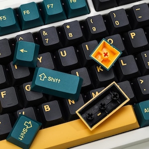 Puede incluir: Primer plano de un teclado mecánico con teclas negras y verde azulado. Las teclas están dispuestas sobre una base blanca. Se han quitado varias teclas, revelando los componentes internos. La tecla "Shift" es visible, junto con otras teclas de función.
