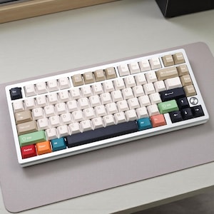 May include: A white and silver mechanical keyboard with a variety of keycap colors, including beige, black, green, and red. The keyboard is on a gray surface, and the keys are arranged in a standard QWERTY layout.