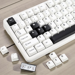 May include: A white mechanical keyboard with black and white keycaps. The keyboard has a compact layout with the letters WASD highlighted in black. Several keycaps are removed, showing the internal components. The keyboard sits on a light wood surface.
