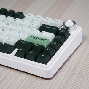 May include: A white mechanical keyboard with a mix of white and dark green keycaps. Some keycaps feature botanical illustrations. The keyboard has a silver knob and is set against a light wood grain surface.