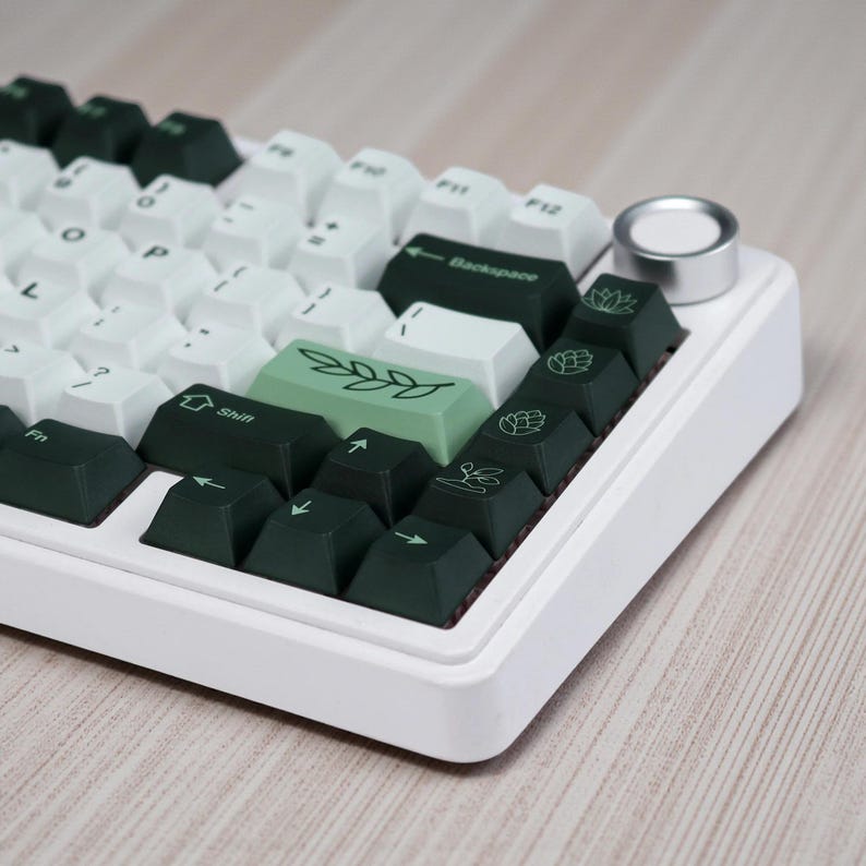 May include: A white mechanical keyboard with a mix of white and dark green keycaps. Some keycaps feature botanical illustrations. The keyboard has a silver knob and is set against a light wood grain surface.