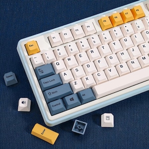 Könnte beinhalten: Eine hellblaue und weiße mechanische Tastatur mit verschiedenen Tastenkappen. Die Tastatur hat weiße und blaue Tastenkappen, einschließlich der "Esc", "Enter" und Funktionstasten in Gelb. Die Tastatur befindet sich auf einer dunkelblauen Oberfläche.