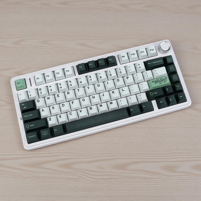 May include: A white mechanical keyboard with black and white keycaps. The keyboard has a silver dial and a green escape key. The keycaps have a botanical design on the space bar and other keys.