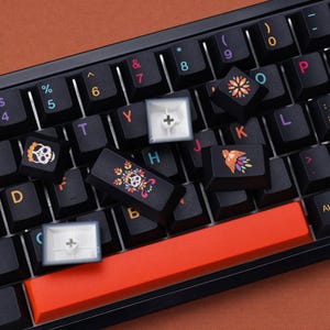 May include: A black mechanical keyboard with custom keycaps. Some keycaps feature colorful Day of the Dead skull and floral designs. The space bar is bright orange. Other keys are black with white lettering and symbols.