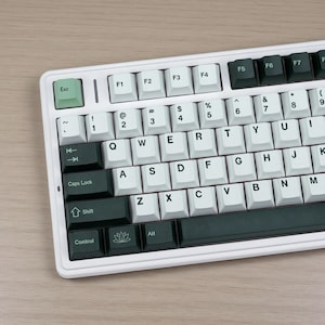 May include: A white mechanical keyboard with a dark green and white keycap set. The keyboard has a standard QWERTY layout with function keys and a number pad. The keycaps are a mix of dark green and white, with the letters and symbols printed in black.