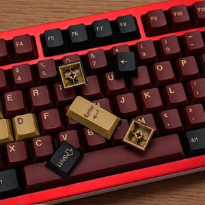 May include: A mechanical keyboard with burgundy keycaps and a red frame. Several keycaps are removed, showing the internal mechanisms. The "Enter" key is gold, and the "Shift" and arrow keys are black.