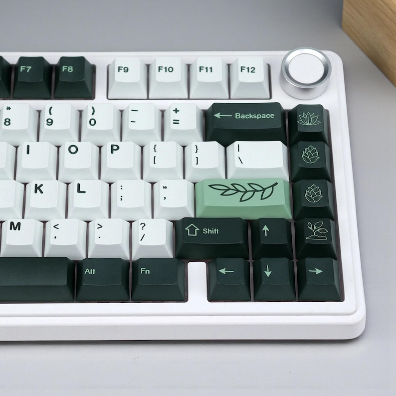 May include: A white mechanical keyboard with dark green and white keycaps. The keycaps feature various symbols and text, including the Backspace key, arrow keys, and function keys. The keyboard also has a silver volume knob.